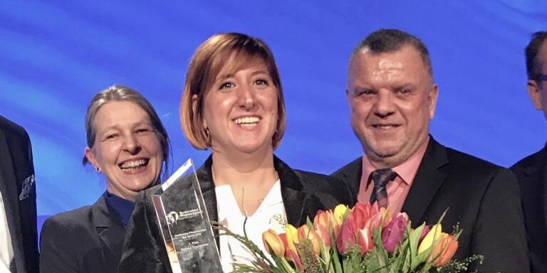 Eine Pflegerin steht auf der Bühne bei einer Preisverleihung mit einem Preis in der einen Hand und einen Blumenstrauß in der anderen. Hinter ihr stehen einige Leute. 